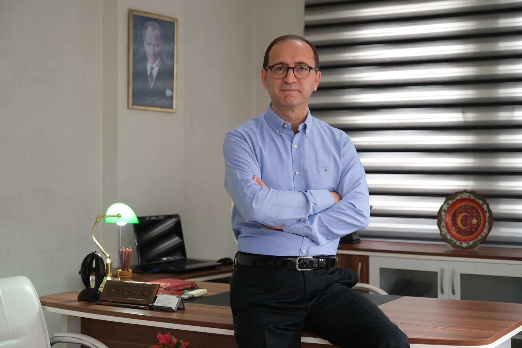 Mustafa Şahin ofisinde, açık mavi gömlekle çalışma masasına yaslanmış portre fotoğrafı